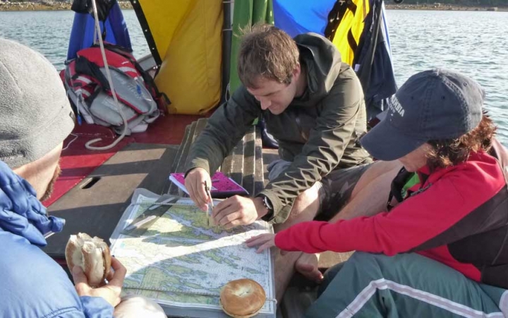 Three people examine a map while on a sailboat. One person eats a bagel, while another bagel rests on the map.